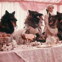 5 dogs, mouths open, dressed in fancy clothes at a fancy dinner table with two footmen behind them.