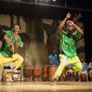 2 dancers wearing green shirts with stars and yellow pants. the dancers are barefoot and smiling. a row of african drummers are behind them.