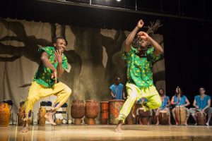 2 dancers wearing green shirts with stars and yellow pants. the dancers are barefoot and smiling. a row of african drummers are behind them.